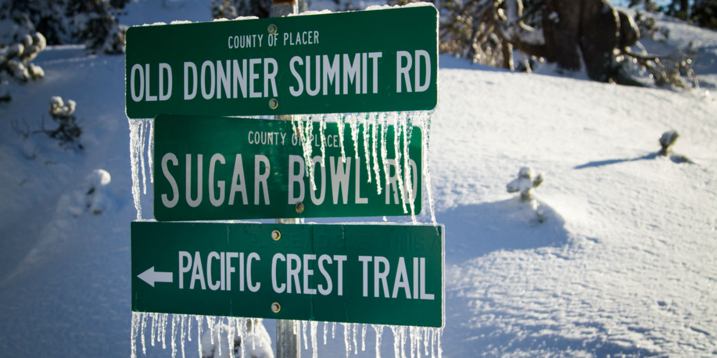 3 frozen green road signs with icicles hanging down. Old Donner Summit Road on top Sugar Bowl Road in the middle and a sign pointing to the Pacific Crest Trail on the bottom.