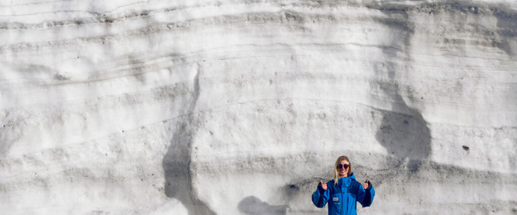 A young woman dwarfed by an enormous wall of snow behind her.