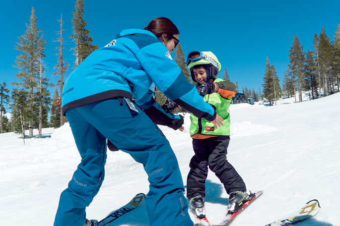 Friendly ski instructor teaching a young boy with a green helmet cover to ski.